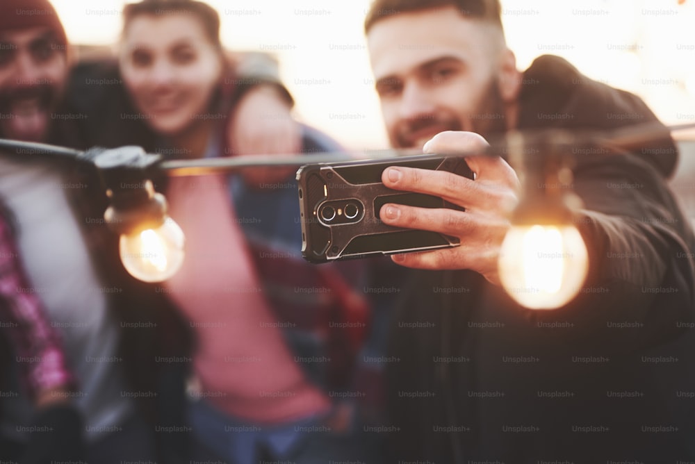 Focused picture. Group of young cheerful friends having fun, hug each other and takes selfie on the roof with decorate light bulbs.