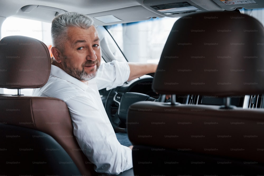 Conception of success. Joyful bearded man in white shirt looks into the camera while sitting in the modern car.