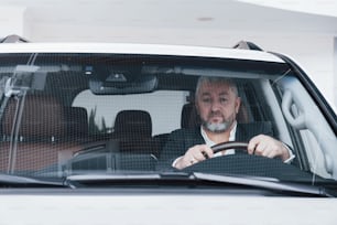 Guarda nella telecamera. Vista frontale dell'uomo d'affari anziano nella sua nuova auto moderna che testa nuove funzioni.