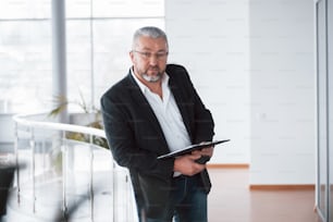 Modern looking place. Photo of senior businessman in the spacious room with plants behind. Holding and reading documents.