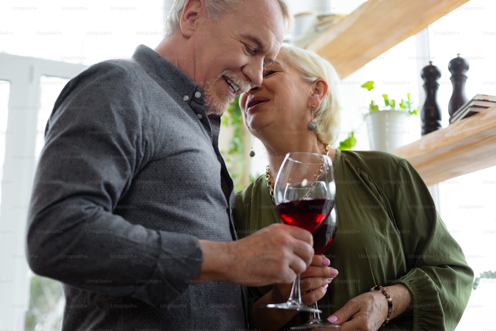 Celebrating with wine. Charming delightful appealing bewitching silver-haired bewitching woman whispering on ear to her elderly grey-haired well-dressed attractive spouse with a glass of wine.