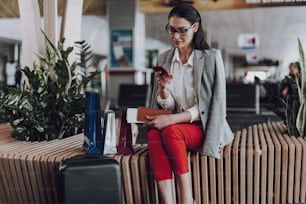 Focus on calm elegant female in glasses sitting on bench in lounge. She is messaging on smartphone. Young lady is waiting for departure with luggage