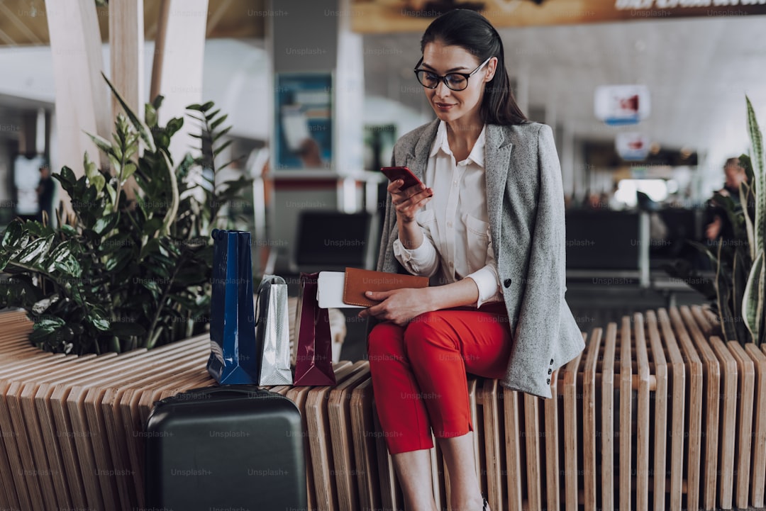 Focus on calm elegant female in glasses sitting on bench in lounge. She is messaging on smartphone. Young lady is waitin