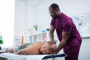 Massage therapist. Dark-skinned massage therapist in uniform and glasses massaging neck for sportsman