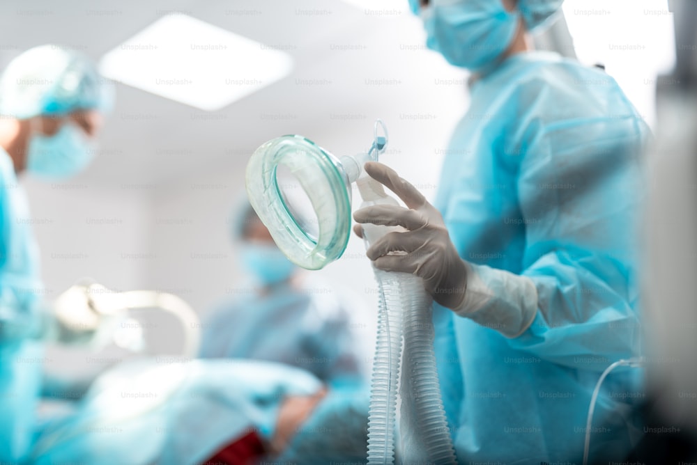 Primer plano del brazo femenino en guante estéril preparando el equipo respiratorio para el paciente durante la cirugía. Señora con máscara protectora y vestido azul