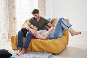 Lying on the boyfriend's legs. Happy couple relaxing on the yellow sofa in the living room of their new house.