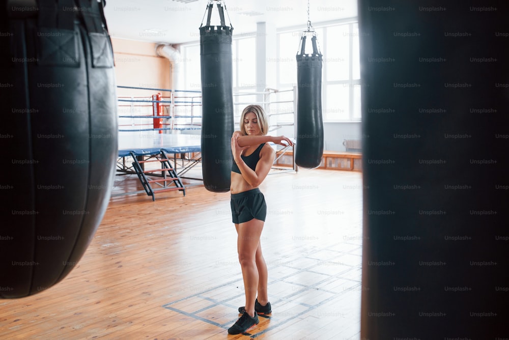 Alone in the room. Woman near the black punching bag in the gym. Natural lighting.