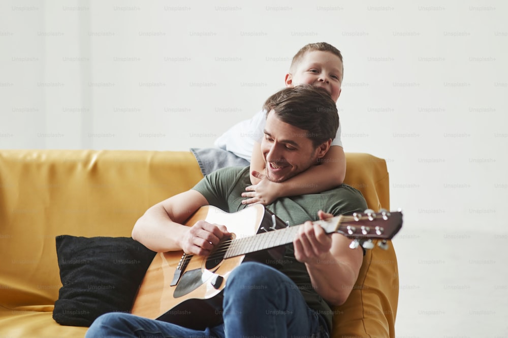 Playful mood. Father and his son sitting on the sofa together. Acoustic guitar in hands.