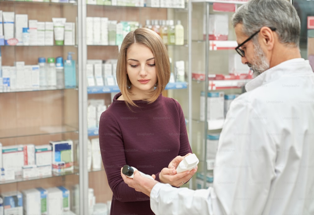 Mature pharmacist offering to customer medical products in drugstore. Beautiful woman holding white cosmetic bottles, choosing. Chemist having grey hair, wearing glasses and white lab coat.