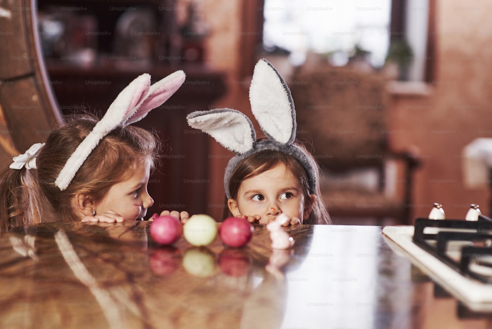 Cute kids. Funny two sisters in bunny ears hidding near the table with painted eggs.