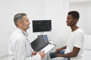 Médico sentado frente al paciente y consultando. Hombre africano con camiseta blanca en examen en gabinete médico, equipado con computadora y pantalla. Terapeuta sosteniendo una carpeta negra.