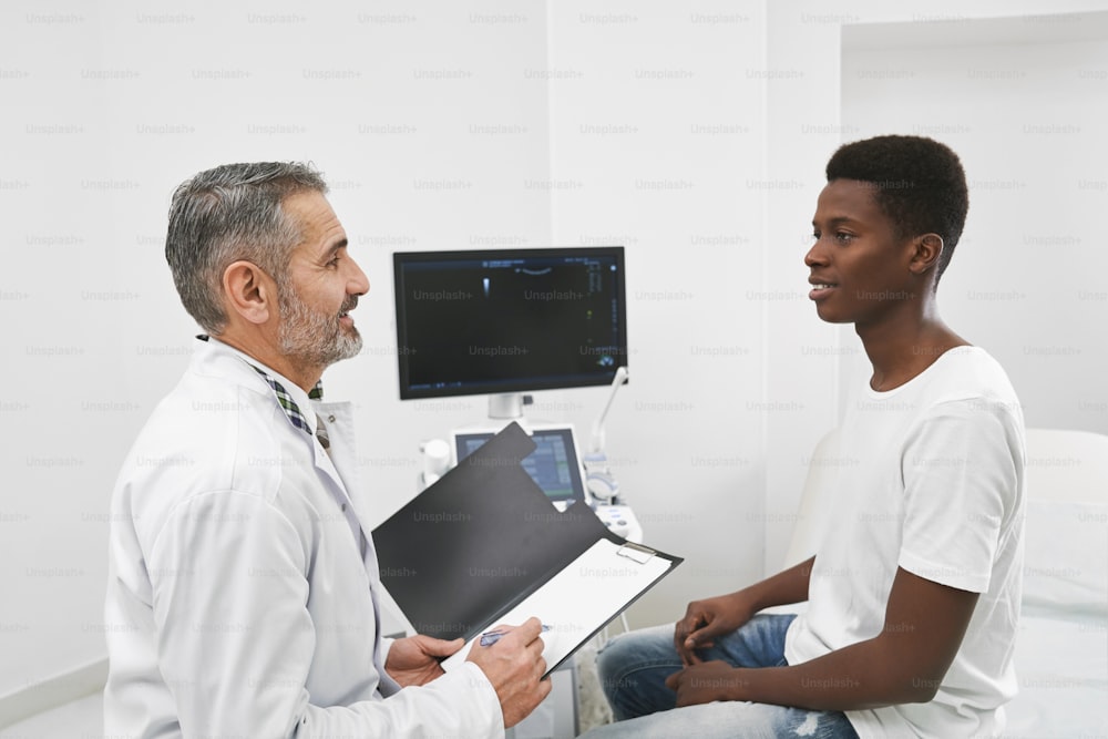 Doctor sitting opposite patient and consulting. African man in white t shirt on examination in medical cabinet, equipped with computer and screen. Therapist holding black folder.