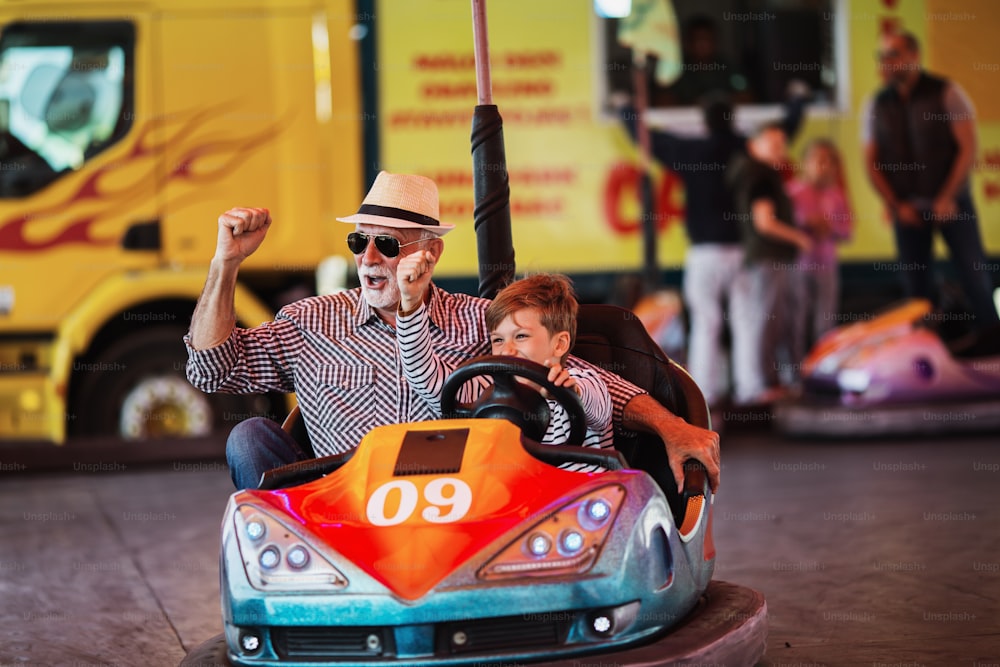 Grandfather and grandson having fun and spending good quality time together in amusement park. They enjoying and smiling while driving bumper car together.