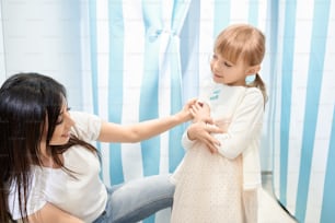 Smiling girl trying on new lovely dress in fitting room. Little daughter shopping together with beautiful mother, choosing and buying outfit in stores. Concept of shopping time and dream.