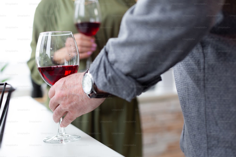 레드 와인 한 잔. 손목에 시계를 차고 테이블 위에 서 있는 레드 와인 한 잔을 들고 있는 남성 손의 클로즈업 샷.