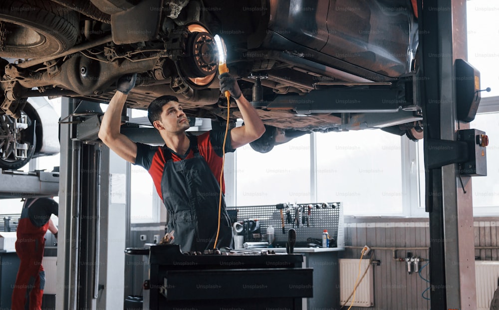 Daylight comes from the windows. Man at the workshop in uniform fixes broken parts of the modern car.