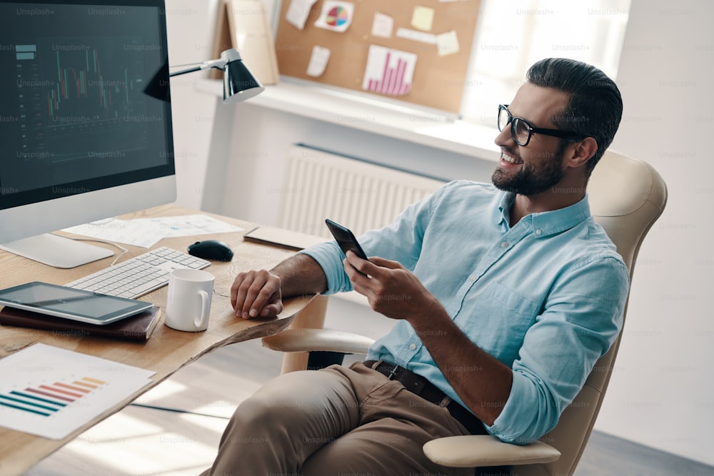 Vista superior do jovem empresário moderno usando o telefone inteligente e sorrindo enquanto sentado no escritório