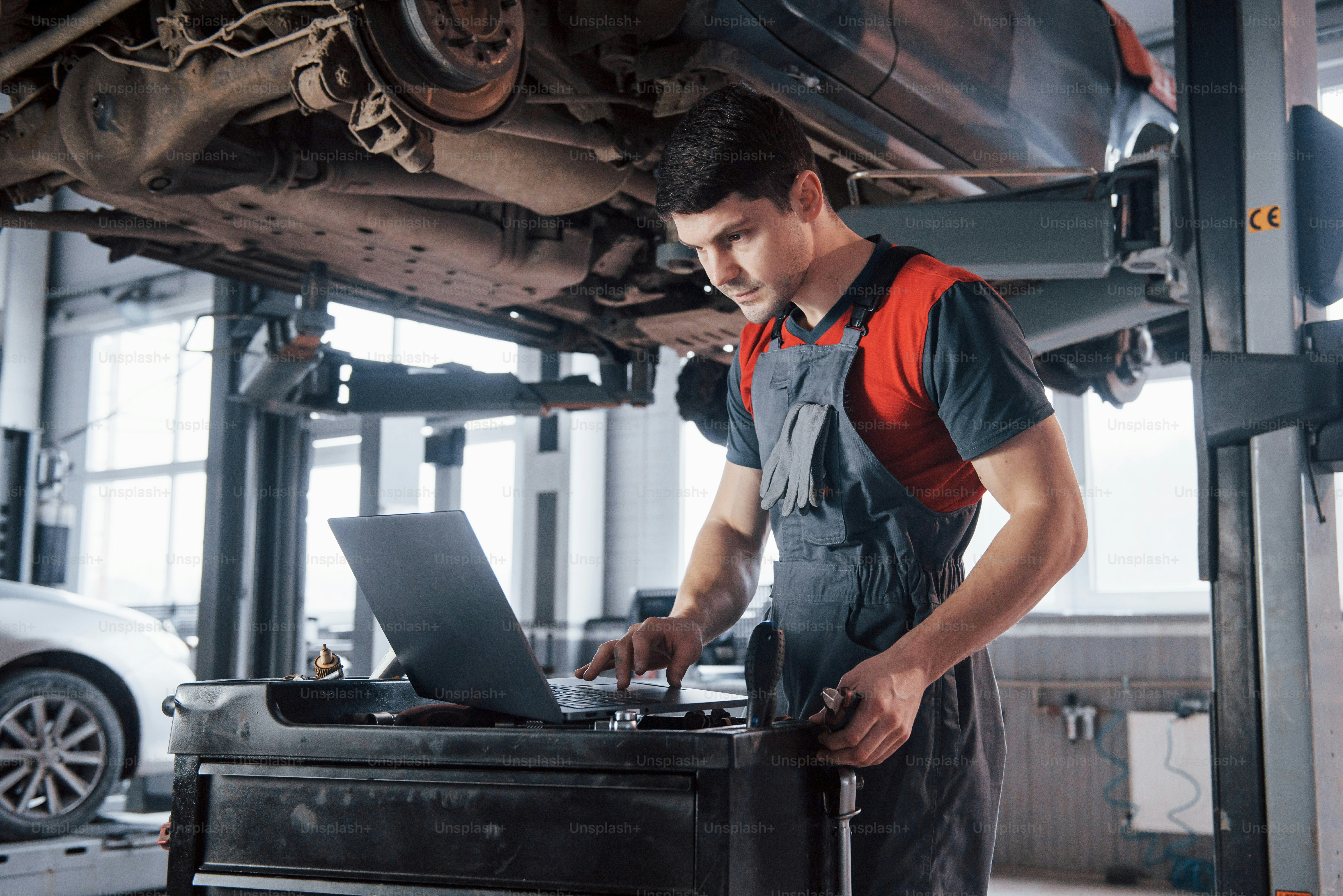 Serious damaged vehicle. Man at the workshop in uniform using laptop for his job for fixing broken car.