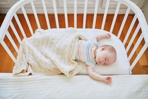 Adorable baby girl sleeping in co-sleeper crib attached to parents' bed. Little child having a day nap in cot. Sleep training concept. Infant kid in sunny nursery