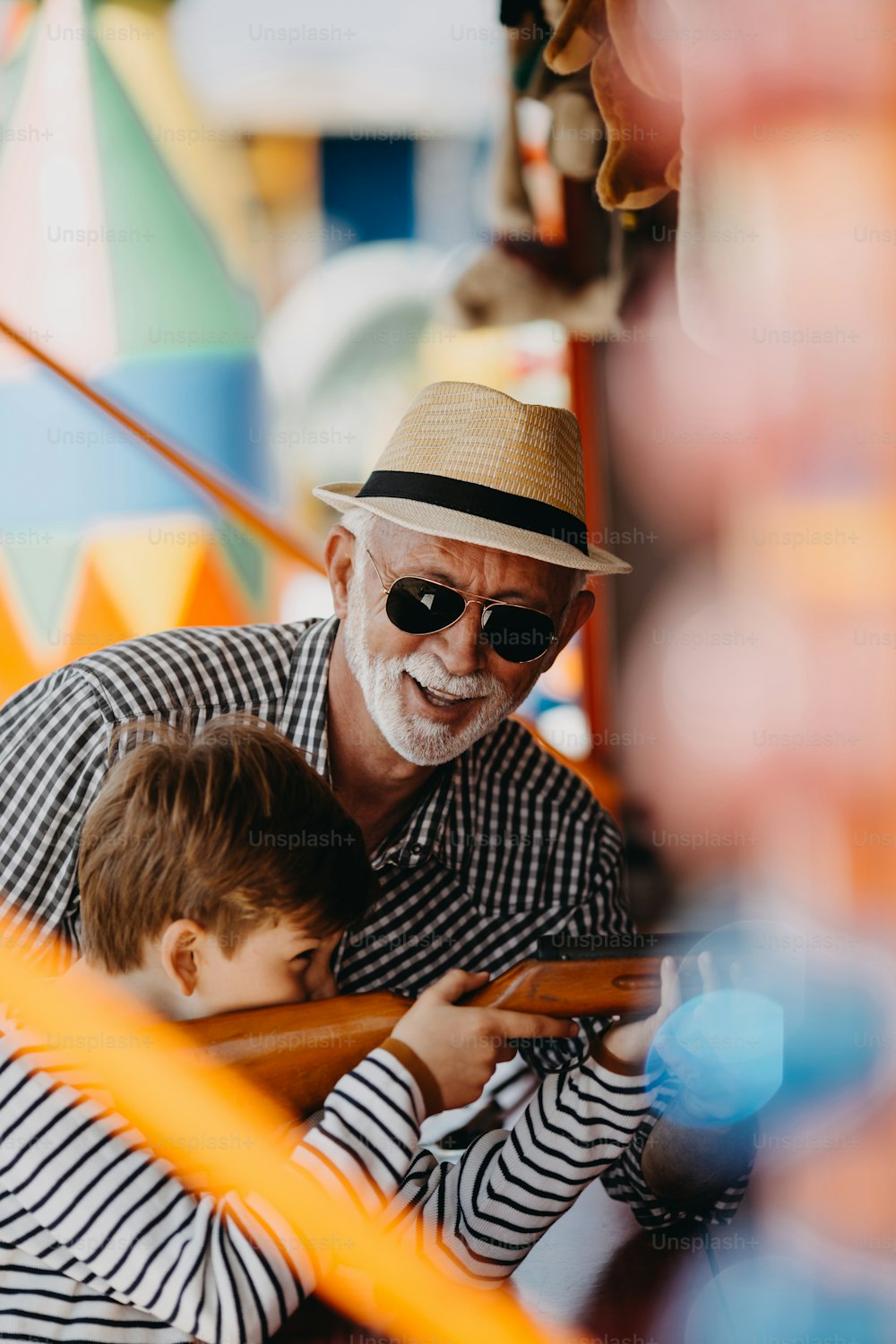 Grandfather and grandson having fun and spending good quality time together in amusement park. Kid shooting with air gun while grandpa helps him to win the prize.