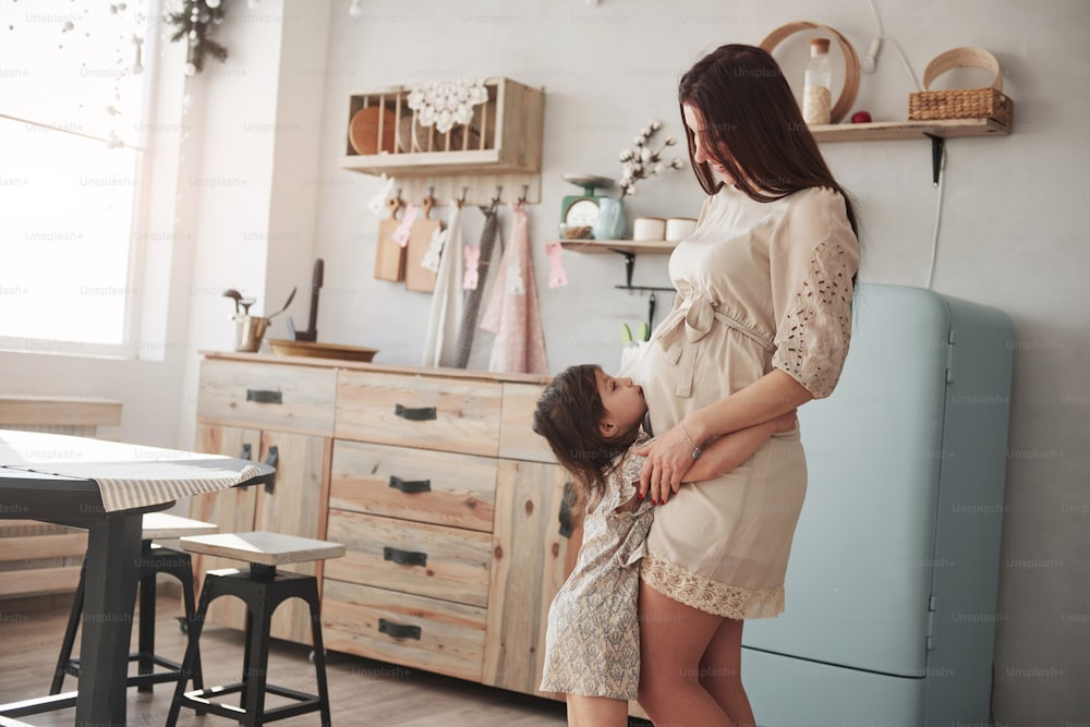 Child kissing stomach. Young positive pregnant brunette stands in the kitchen at daytime.