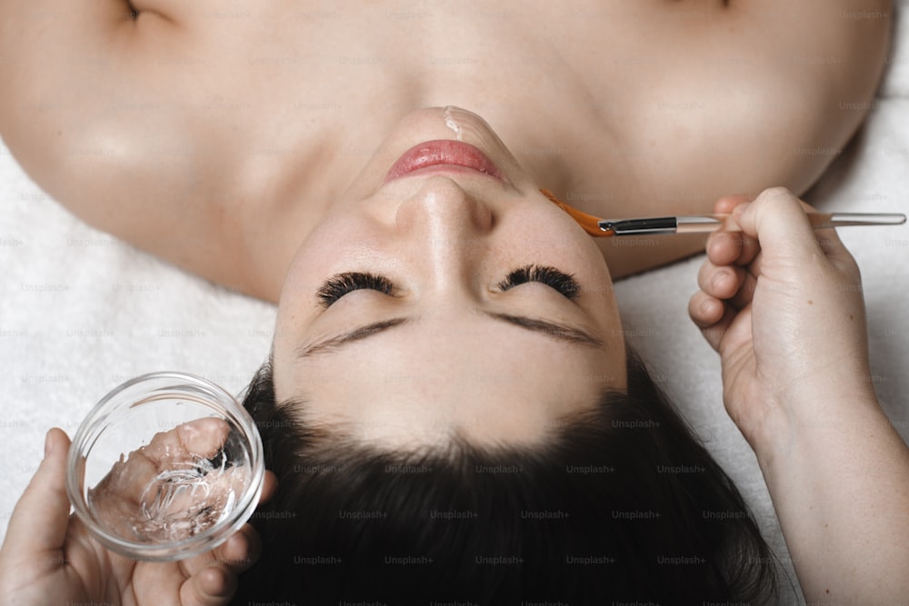 Oberes Nahaufnahmeporträt einer gutaussehenden jungen kaukasischen Frau, die sich mit geschlossenen Augen auf ein Spa-Bett stützt, während sie eine transparente Gesichtsmaske von Kosmetikerin macht.