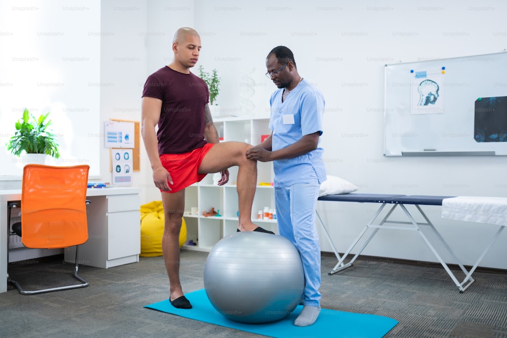 Touching knee. Physical therapist in blue uniform touching knee of sportsman standing near fit ball