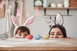 Looks at the eggs. Mother and daughter in bunny ears at easter time have some fun in the kitchen at daytime.