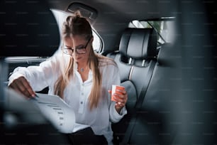 Tazza rossa con caffè nella mano sinistra. L'intelligente donna d'affari siede sul sedile posteriore dell'auto di lusso con interni neri.