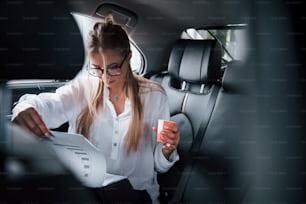 Red cup with coffee in left hand. Smart businesswoman sits at backseat of the luxury car with black interior.