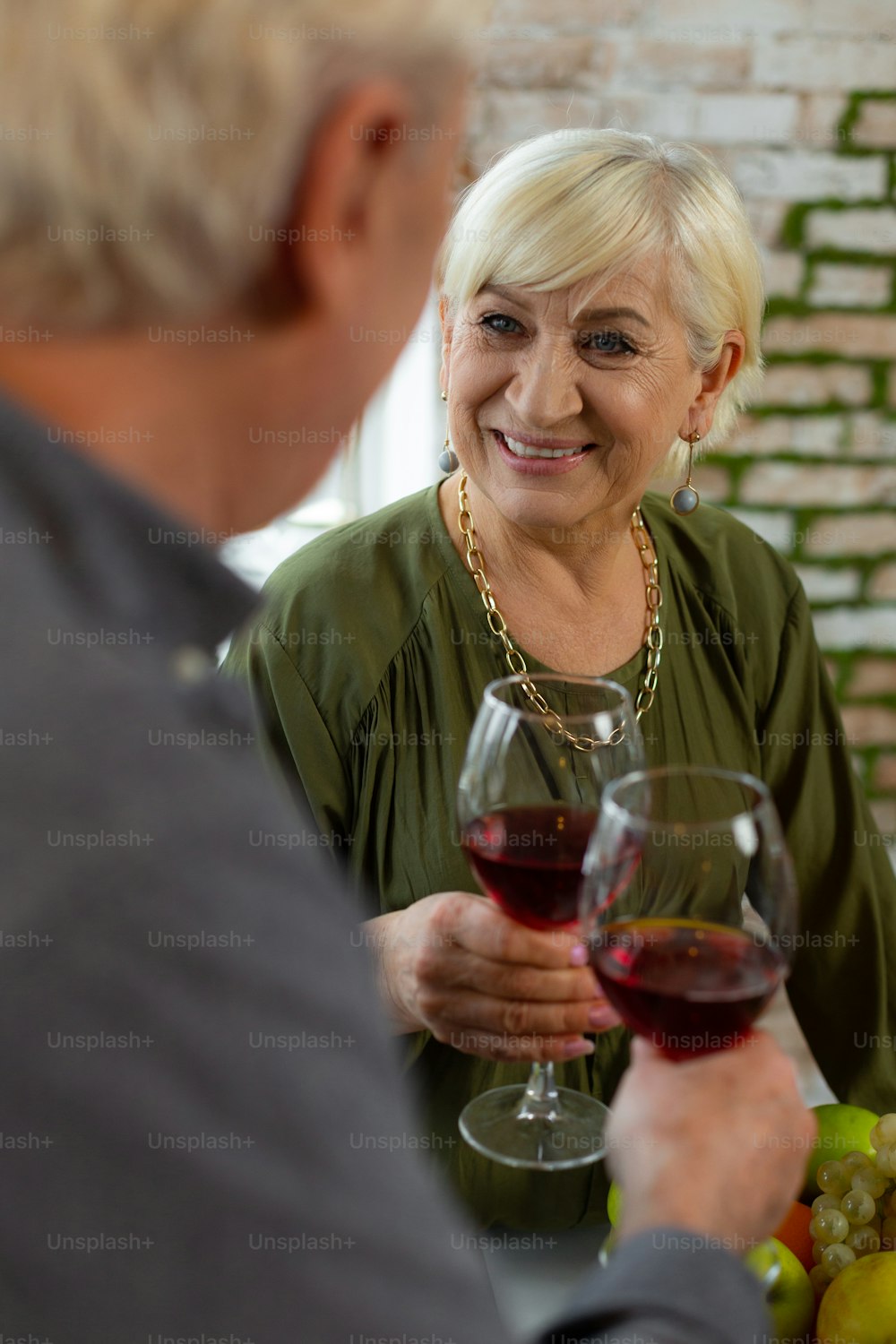 Lovingly look at man. Loving happy smiling beaming glowing white-haired alluring good-looking female wearing emerald elegant garment looking at husband and holding a glass of wine.