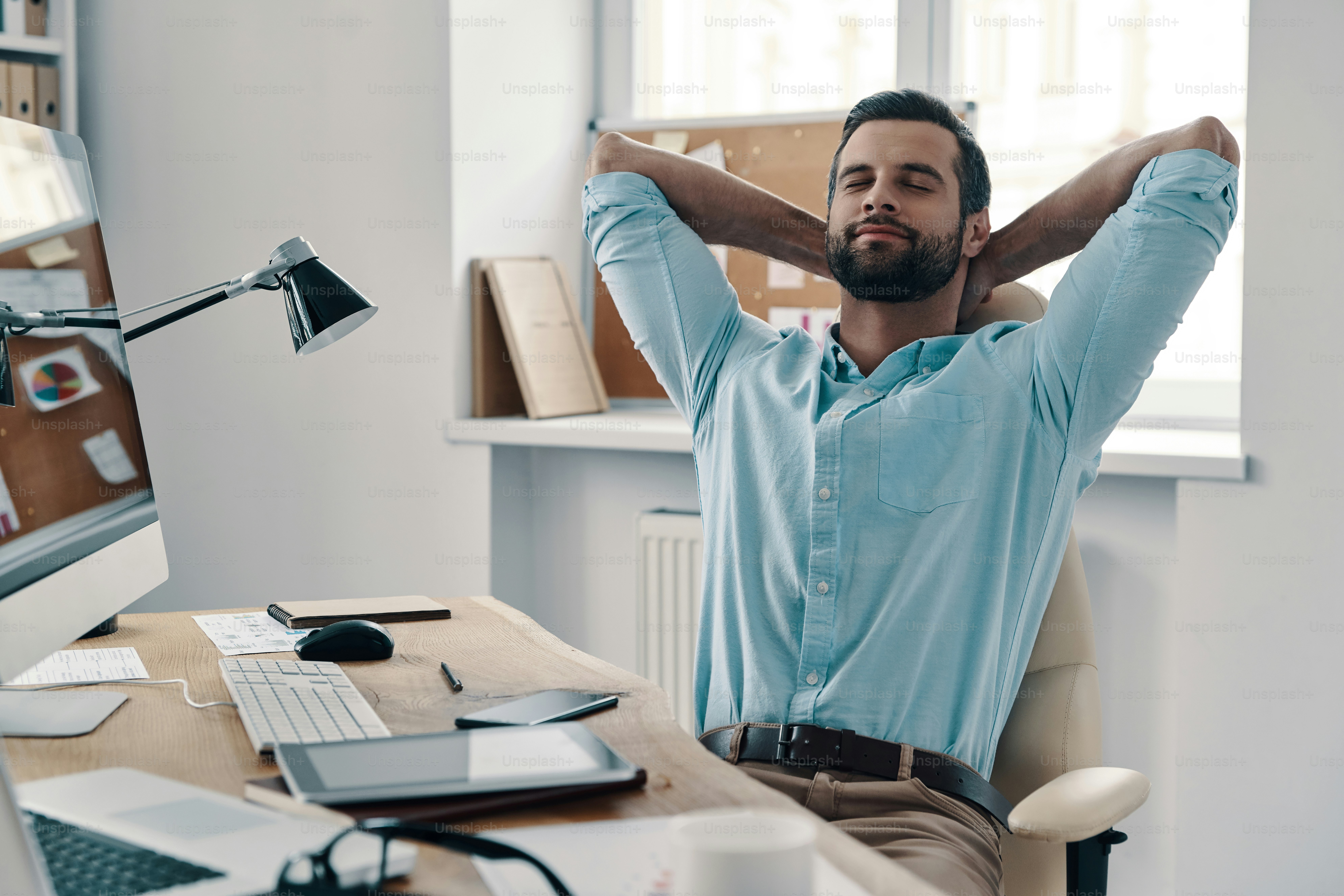 Young modern businessman keeping hands behind head and smiling while sitting in the office
