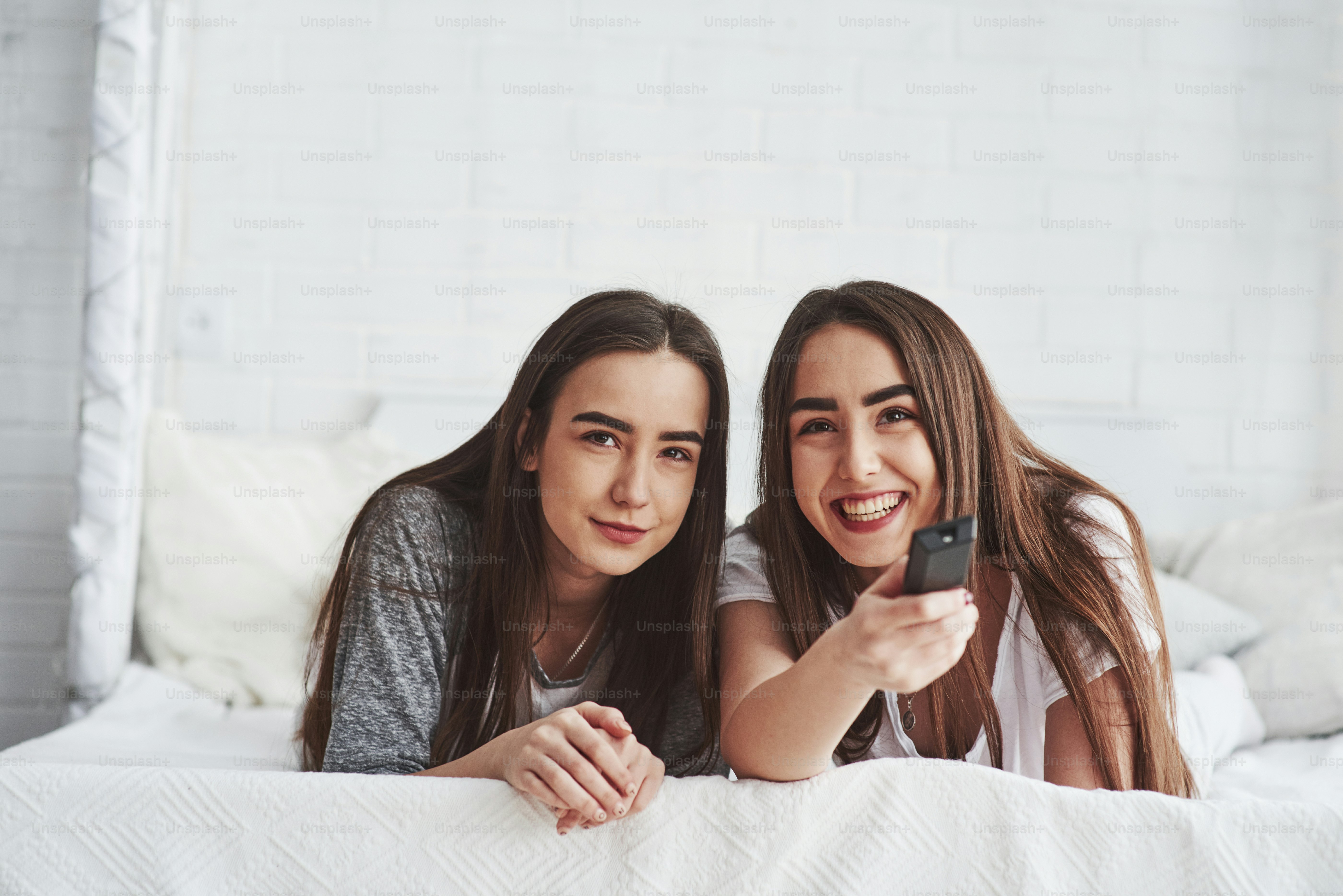 That's moment is so funny, look. Two female twins lying on the white bed at daytime watching movie.