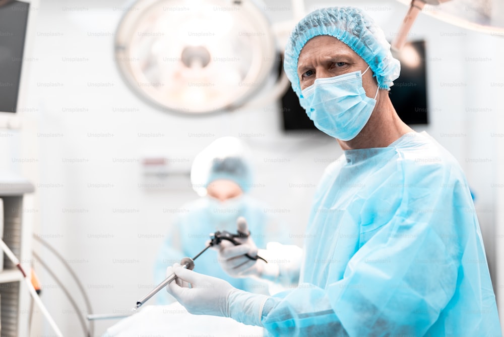 Portrait of middle aged doctor in sterile gloves holding surgical grasper with trocar while looking at camera with serious expression. He is wearing protective mask and blue gown