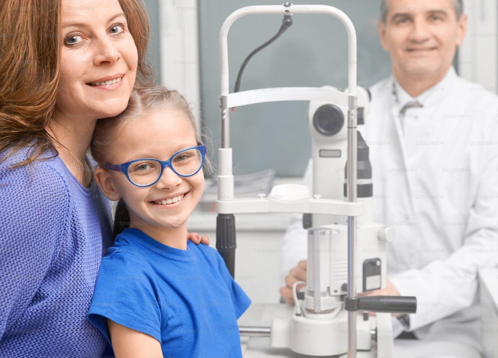 Pretty girl in blue glasses posing in medical ophthalmology clinic with mother. Mature doctor eye specialist smiling, looking at camera, consulting patients.