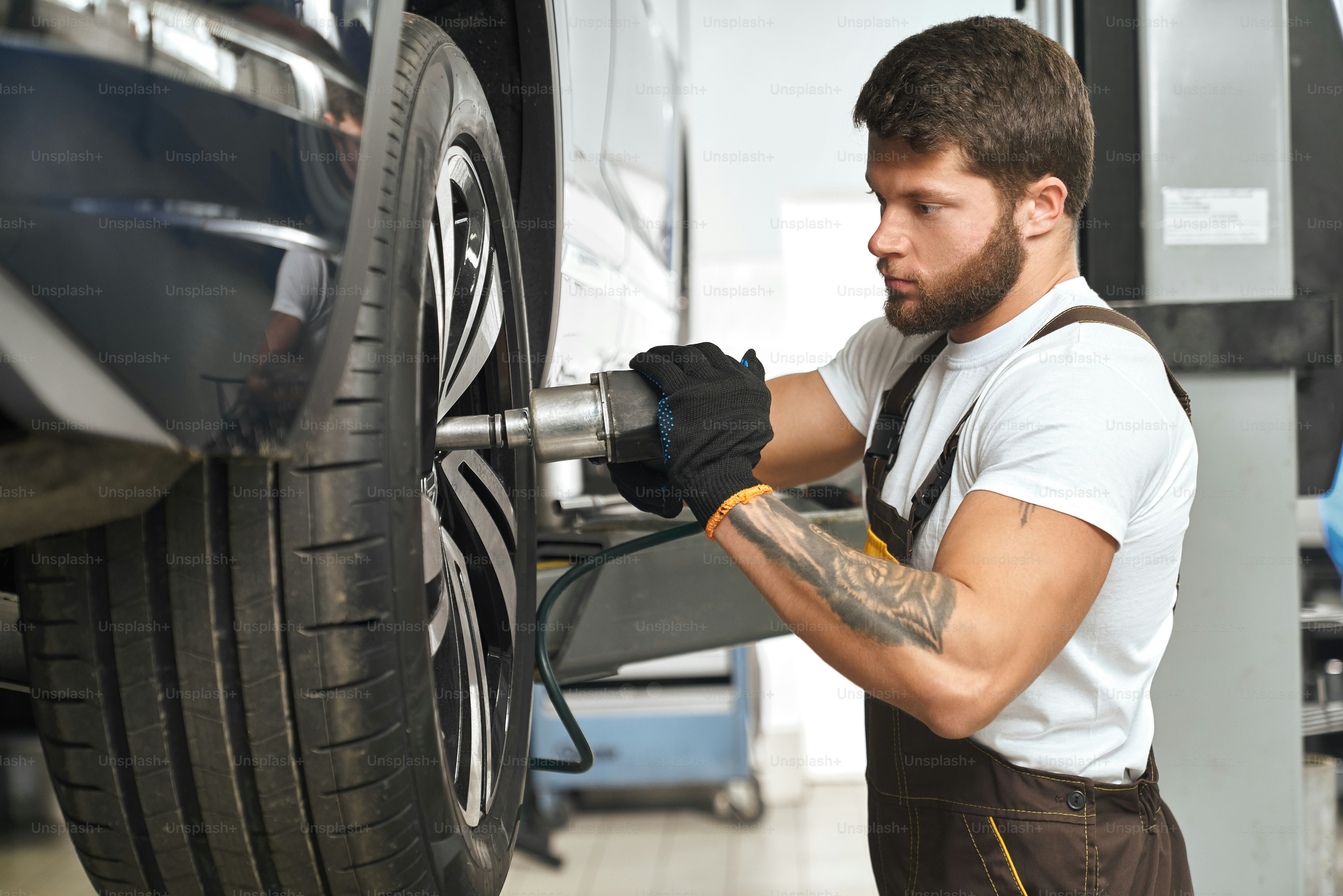 Mudança mecânica séria e concentrada, reparo da calota das rodas, utilizando equipamentos especiais. Homem barbudo e musculoso de camiseta branca consertando veículo na garagem autoservice.