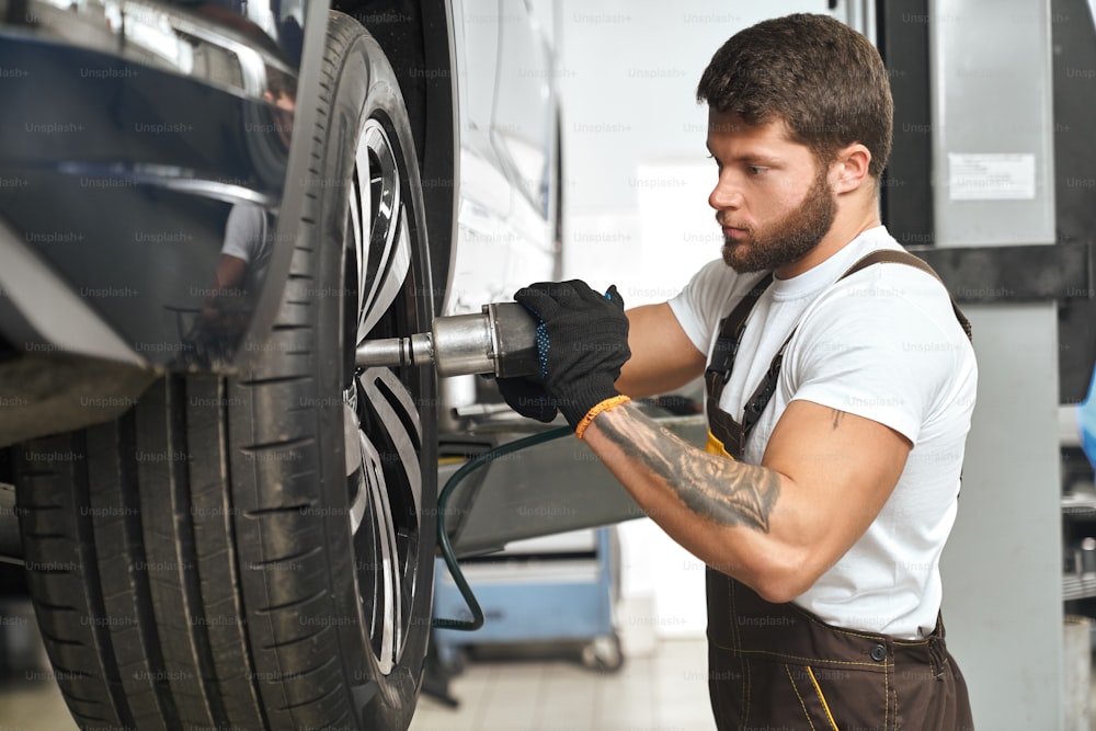 Cambio mecánico serio y concentrado, reparación del tapacubos de la rueda, uso de equipos especiales. Hombre barbudo y musculoso con camiseta blanca arreglando vehículo en garaje autoservice.