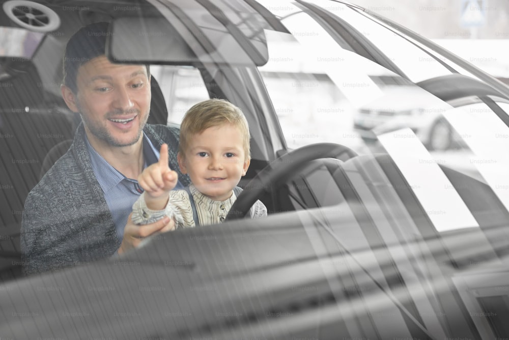 Cute boy sitting with father in new automobile, looking at camera and laughing. Father with child examining auto salon and testing vehicles in showroom. Concept of car presentation.
