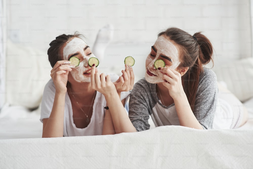 Lying on the whte bed. Conception of skin care by using fresh cucumber rings and white mask on the face. Two female sisters have weekend at bedroom.