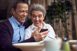 Enjoyable meetings. Waist up portrait of happy smiling aged man embracing woman while they looking at mobile phone and sitting in cafe