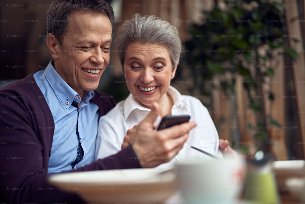 Enjoyable meetings. Waist up portrait of happy smiling aged man embracing woman while they looking at mobile phone and sitting in cafe