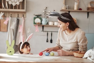Woman shows red egg using forefinger. Mother and daughter in bunny ears at easter time have some fun in the kitchen at daytime.