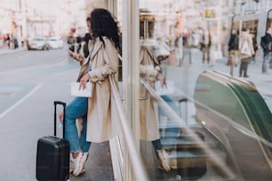 Full length portrait of charming young woman with travel trolley bag reading messages on smartphone