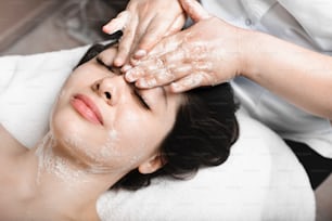 Upper view of a lovely young caucasian female leaning on a spa bed with closed eyes having a facial massage with foam for removing dead skin .