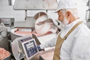 Elderly man worker wearing in white uniform, white cap, brown apron and rubber gloves working on line of manufacturing meat production. Worker standing near equipment for grinding meat.