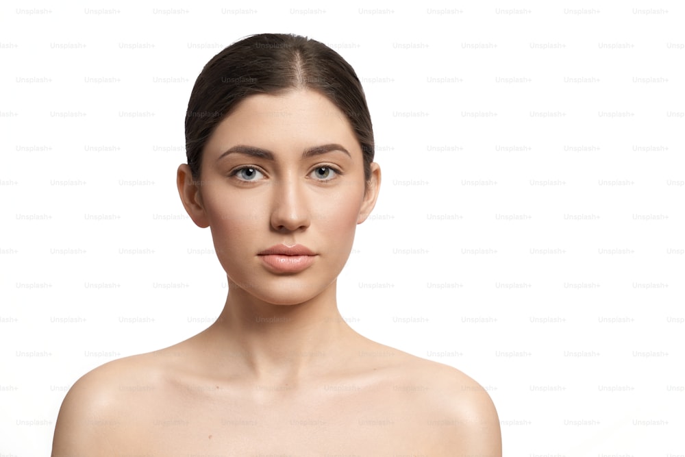 Front view of young woman with plump eyes and dark hair looking at camera on white isolated background. Pretty female with nude makeup posing in studio. Concept of skin care and cosmetology.