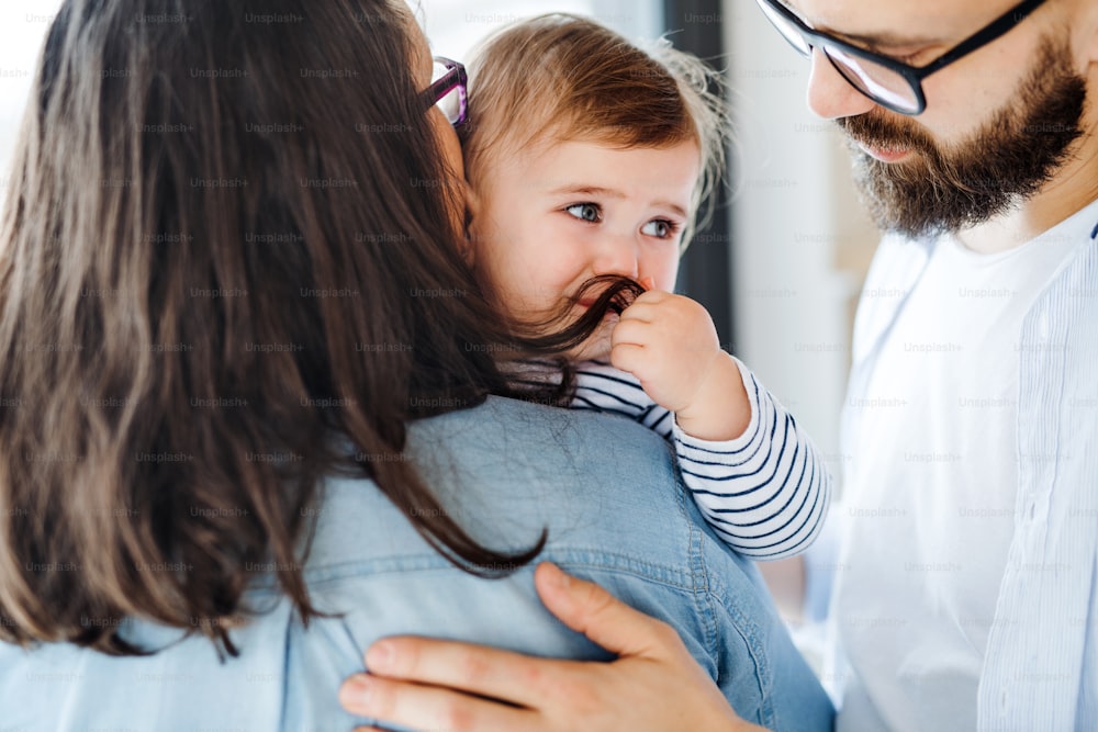 A young family with an unhappy crying toddler girl standing indoors at home, midsection.