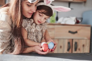 Holding painted eggs. Mother and daughter in bunny ears at easter time have some fun in the kitchen at daytime.