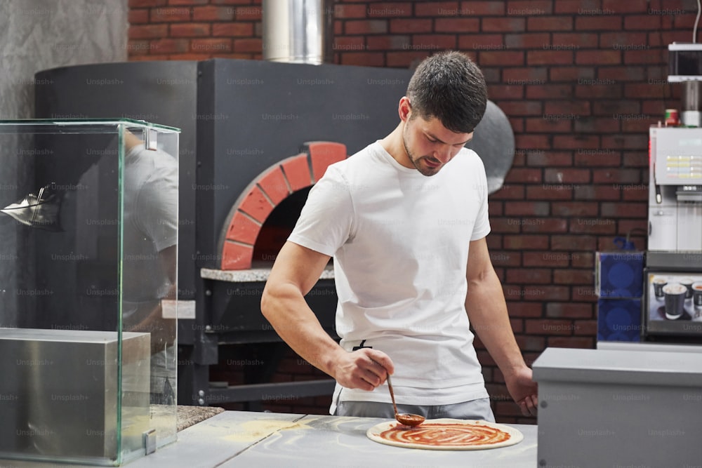 Le poêle est derrière. Boulanger en chemise blanche mettant de la sauce pour faire de délicieuses pizzas pour une commande au restaurant.