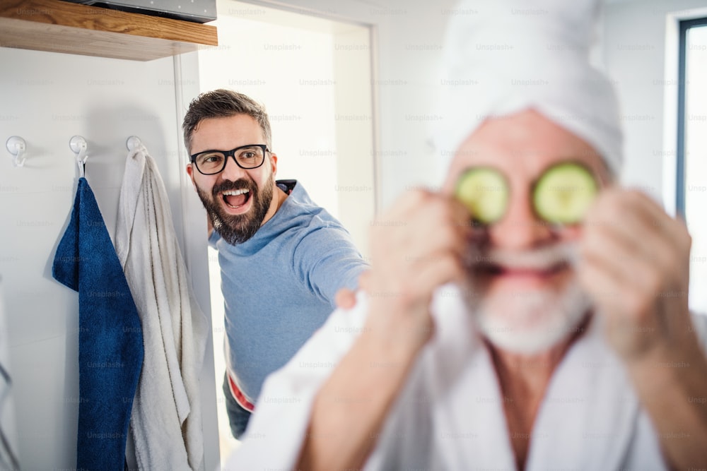 A cheerful adult hipster son and senior father in bathroom indoors at home, having fun.